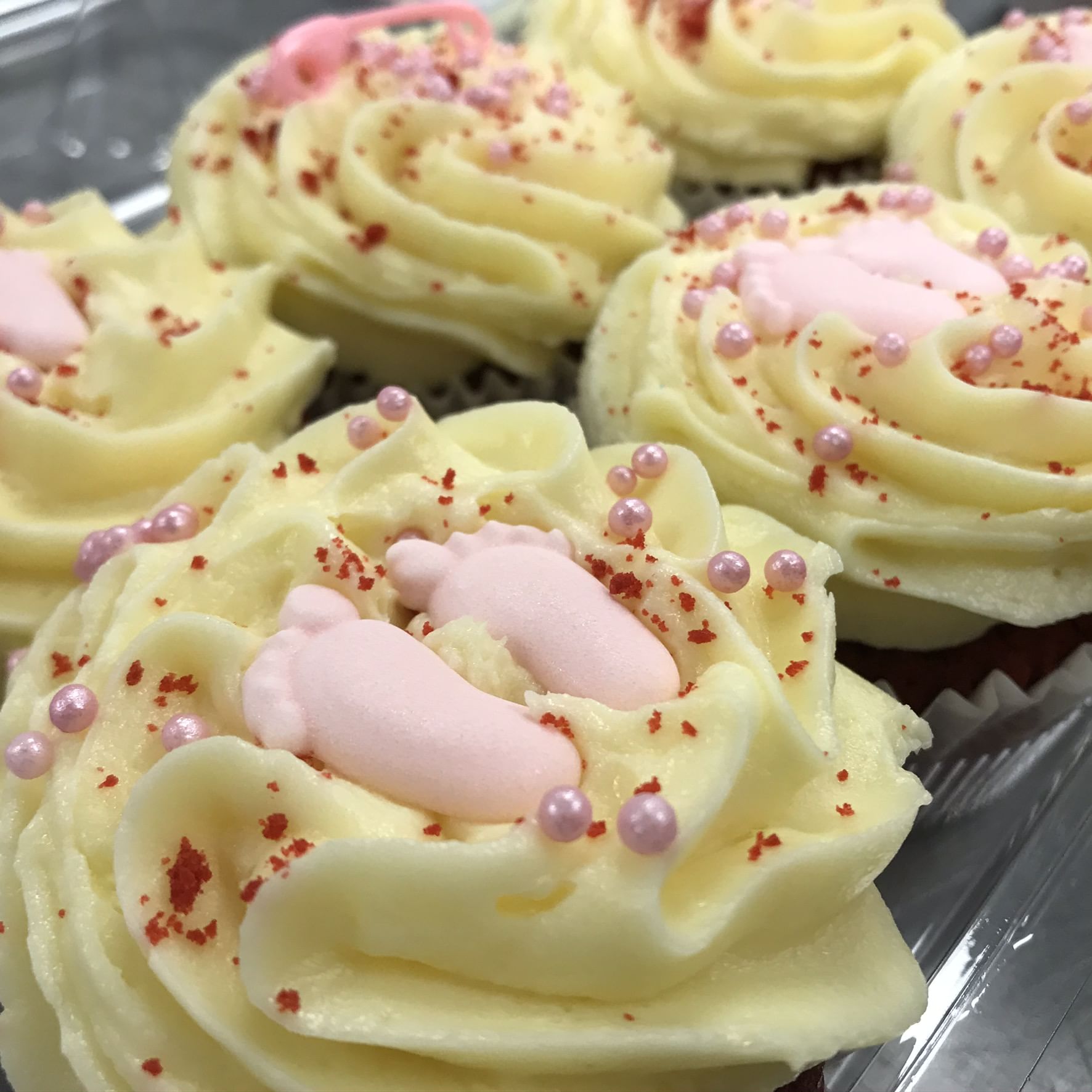 Close-up of red velvet cupcakes topped with creamy yellow frosting, pink baby feet-shaped decorations, and pink candy pearls. Red sprinkles are scattered on the frosting, creating an appealing and festive look. The buttermilk-infused cupcakes are arranged in a bakery display case.